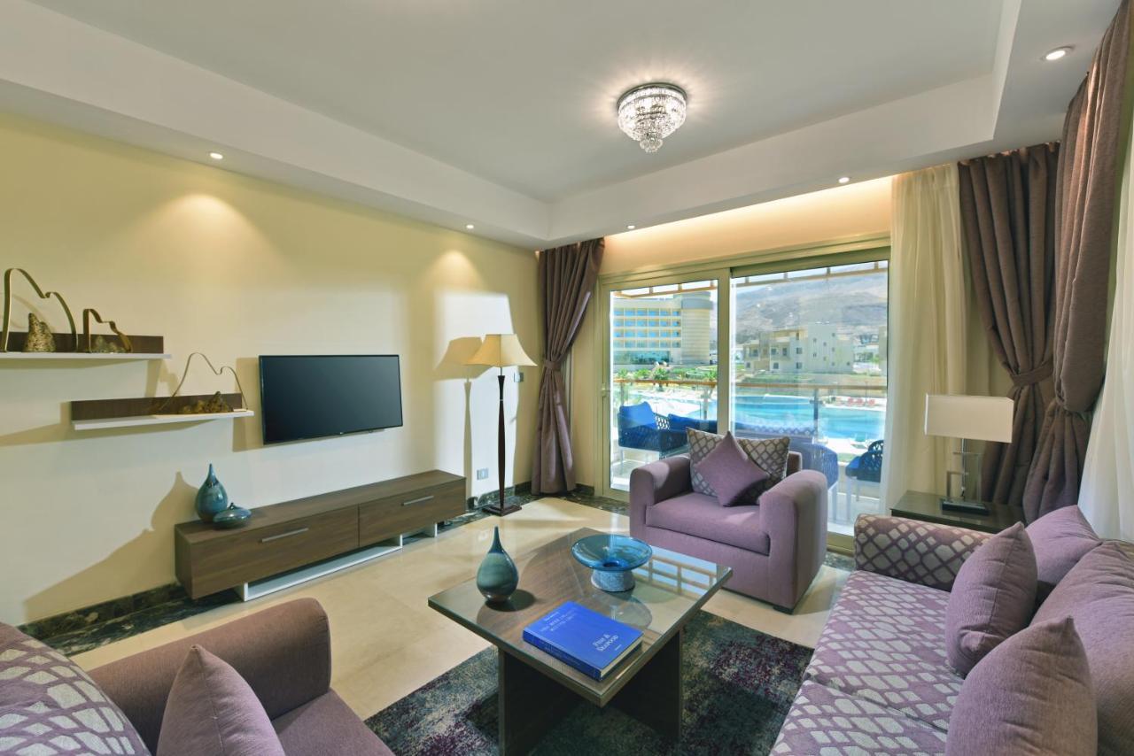 Tolip Resort El Galala Majestic Ain Sukhna Luaran gambar The photo shows a modern living room that features a stylish and cozy design. There are two purple sofas with decorative cushions arranged around a coffee table. The coffee table has a blue book and a decorative item on it. A flat-screen TV is mounte