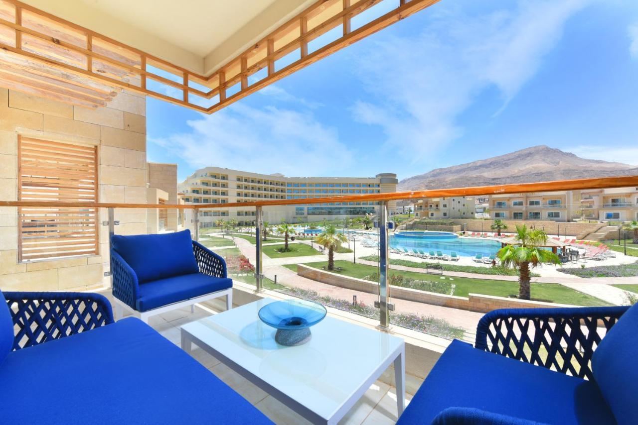 Tolip Resort El Galala Majestic Ain Sukhna Luaran gambar The photo showcases a balcony view featuring two blue chairs and a table with a decorative bowl. The balcony overlooks a landscape that includes a swimming pool and manicured lawns. In the background, there are multiple buildings and palm trees, all 