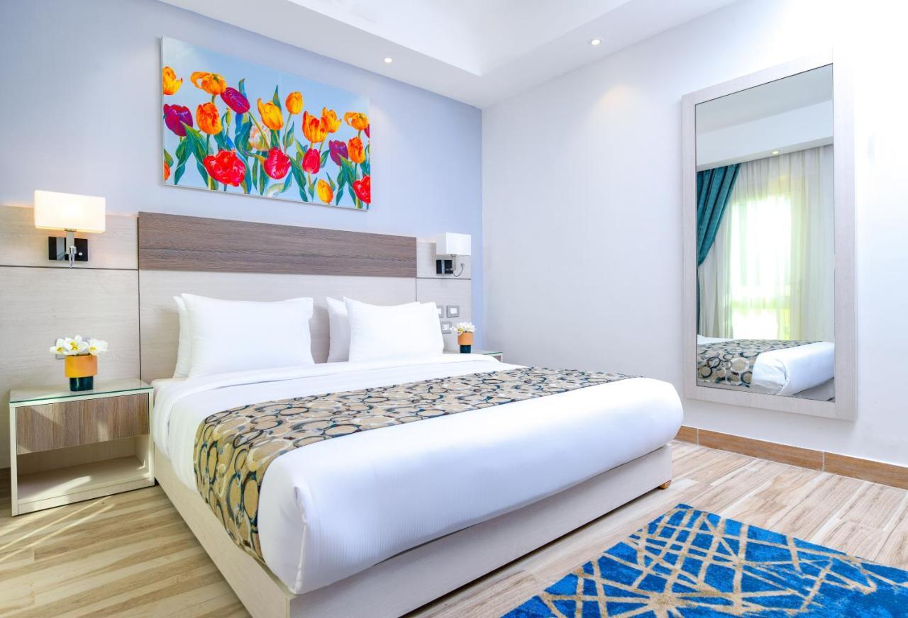 Tolip Resort El Galala Majestic Ain Sukhna Luaran gambar The photo shows a modern hotel bedroom. It features a large bed with white linens and a decorative throw blanket. The room has light wooden flooring and is bright, with ample natural light coming from a window. There is a colorful painting of flowers