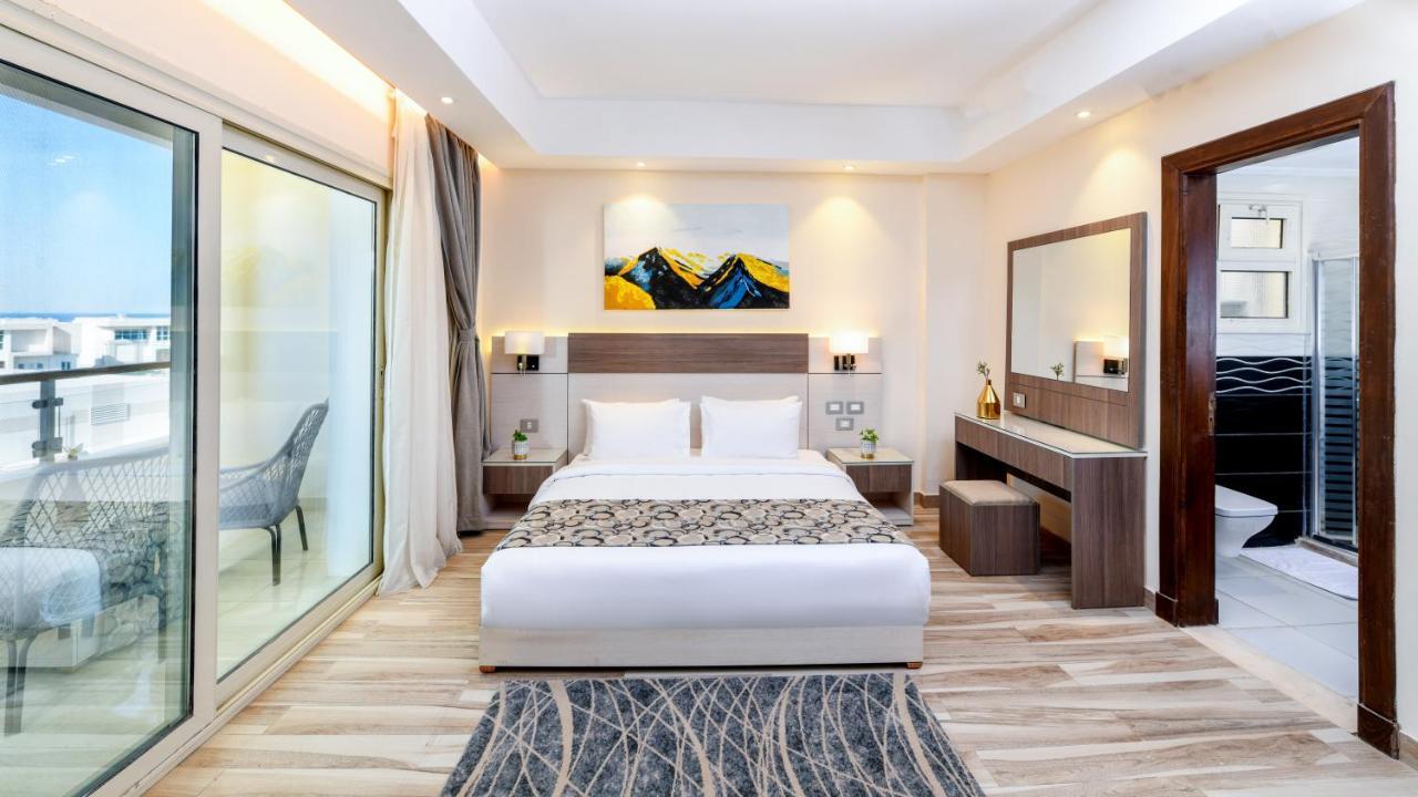 Tolip Resort El Galala Majestic Ain Sukhna Luaran gambar The photo shows a modern hotel room featuring a large bed with white linens and a decorative throw. The room has a neutral color palette, accented with warm lighting. There is a piece of artwork above the bed depicting mountains. To the left, there a