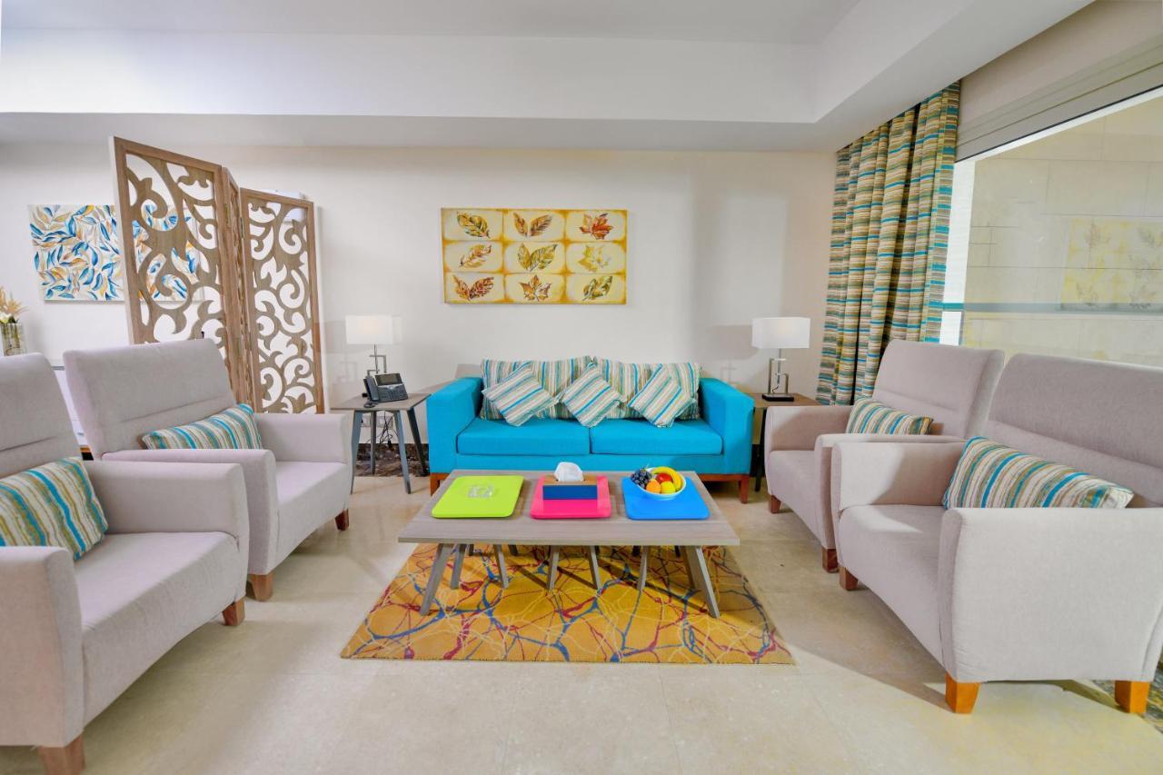 Tolip Resort El Galala Majestic Ain Sukhna Luaran gambar The photo shows a stylish and colorful living room. It features a bright blue sofa with patterned cushions, complemented by two light gray armchairs. In front of the sofa, there's a wooden coffee table with vibrant trays in pink, green, and yellow. A