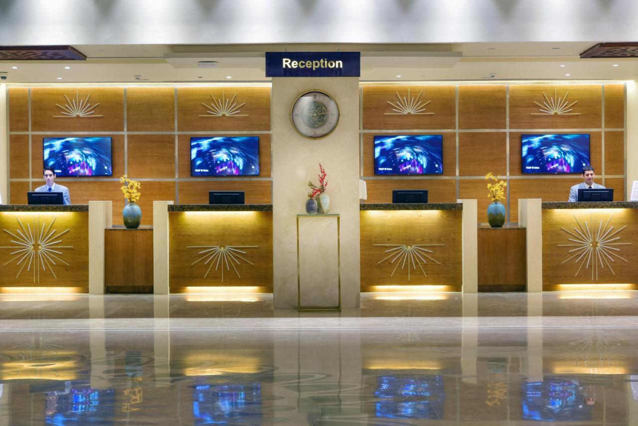 Tolip Resort El Galala Majestic Ain Sukhna Luaran gambar The image shows a hotel reception area. There are several reception desks lined up against a wall with decorative paneling. Each desk has a staff member, and above the desks, there are screens displaying information. A clock is visible on the wall, a