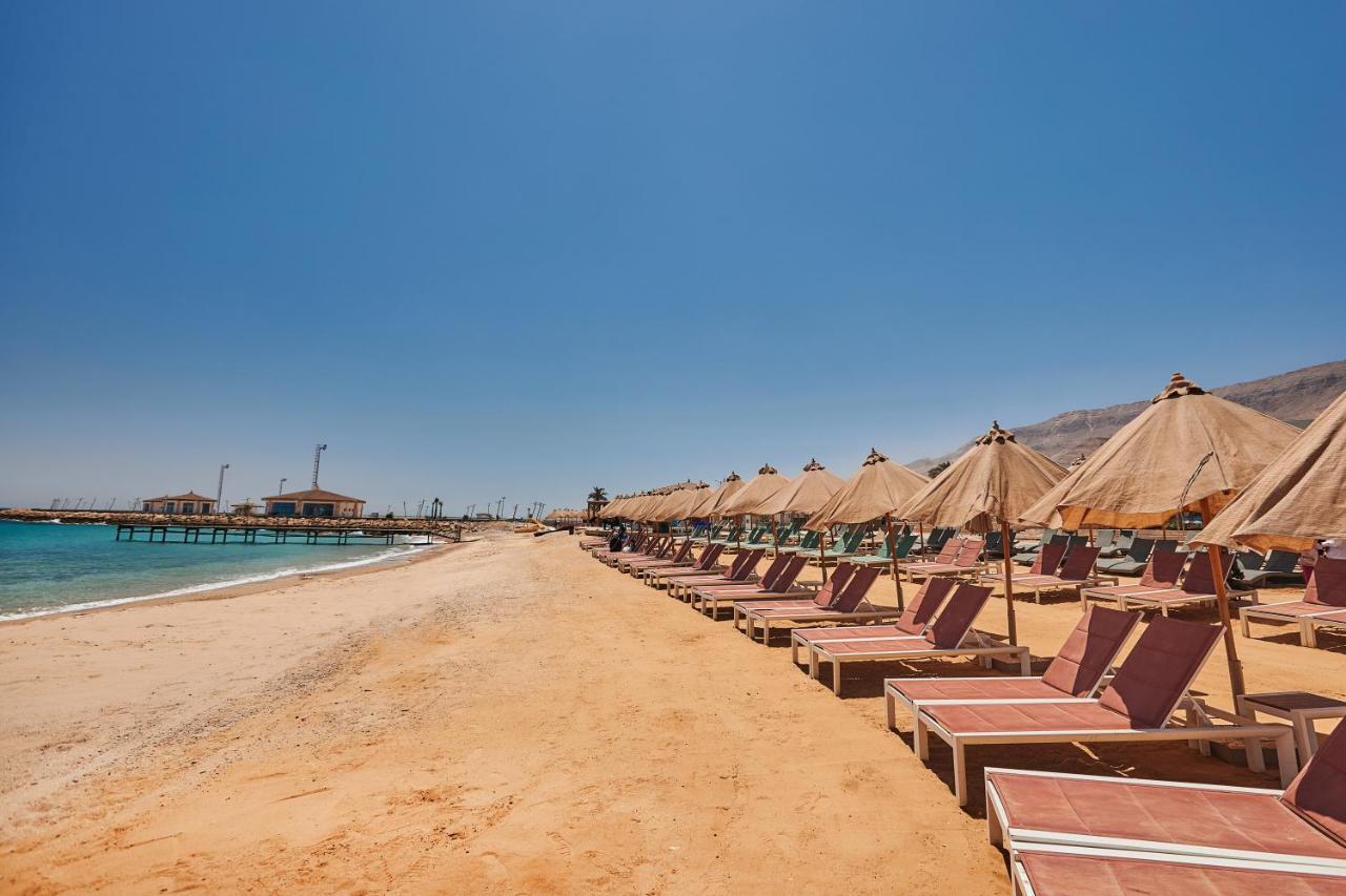 Tolip Resort El Galala Majestic Ain Sukhna Luaran gambar The photo shows a scenic beach with a stretch of golden sand under a clear blue sky. There are several beach loungers arranged in rows, each accompanied by thatched umbrellas providing shade. In the background, there is a wooden pier extending into t