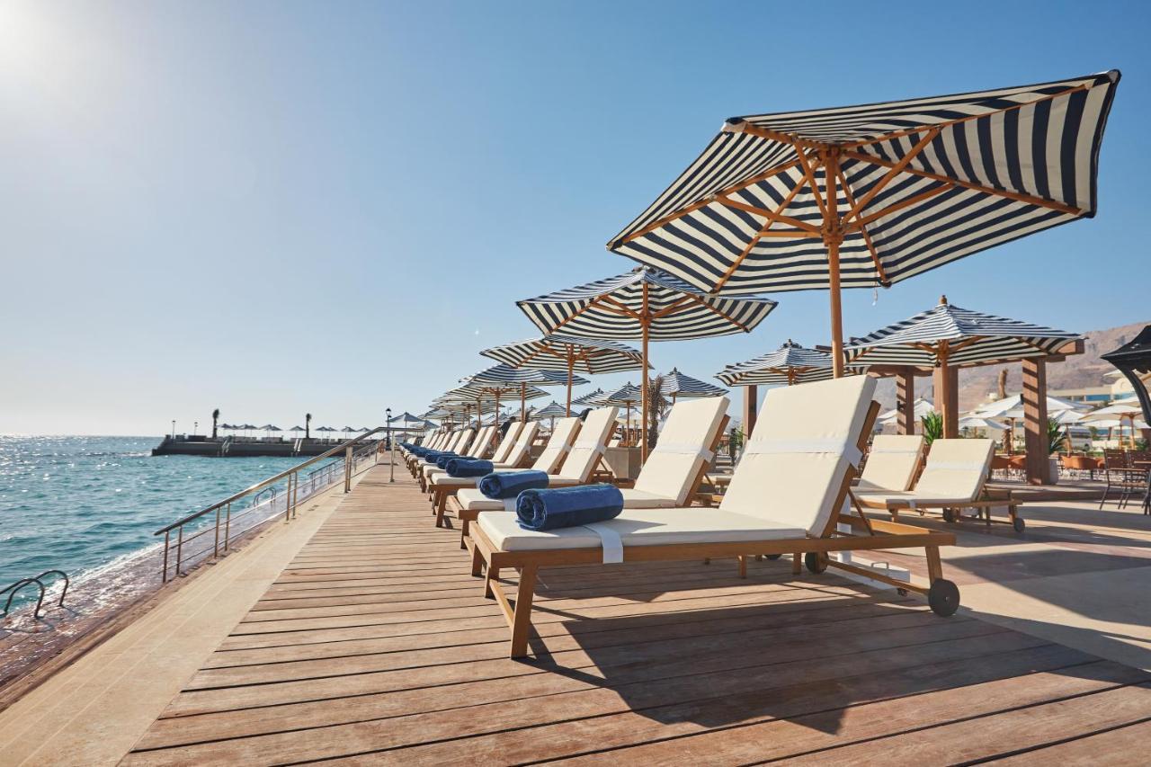 Tolip Resort El Galala Majestic Ain Sukhna Luaran gambar The photo depicts a serene beachside setting featuring a row of sun loungers arranged on a wooden deck. Each lounger is equipped with a folded towel and shaded by striped umbrellas. In the background, the water sparkles under bright sunlight, and the