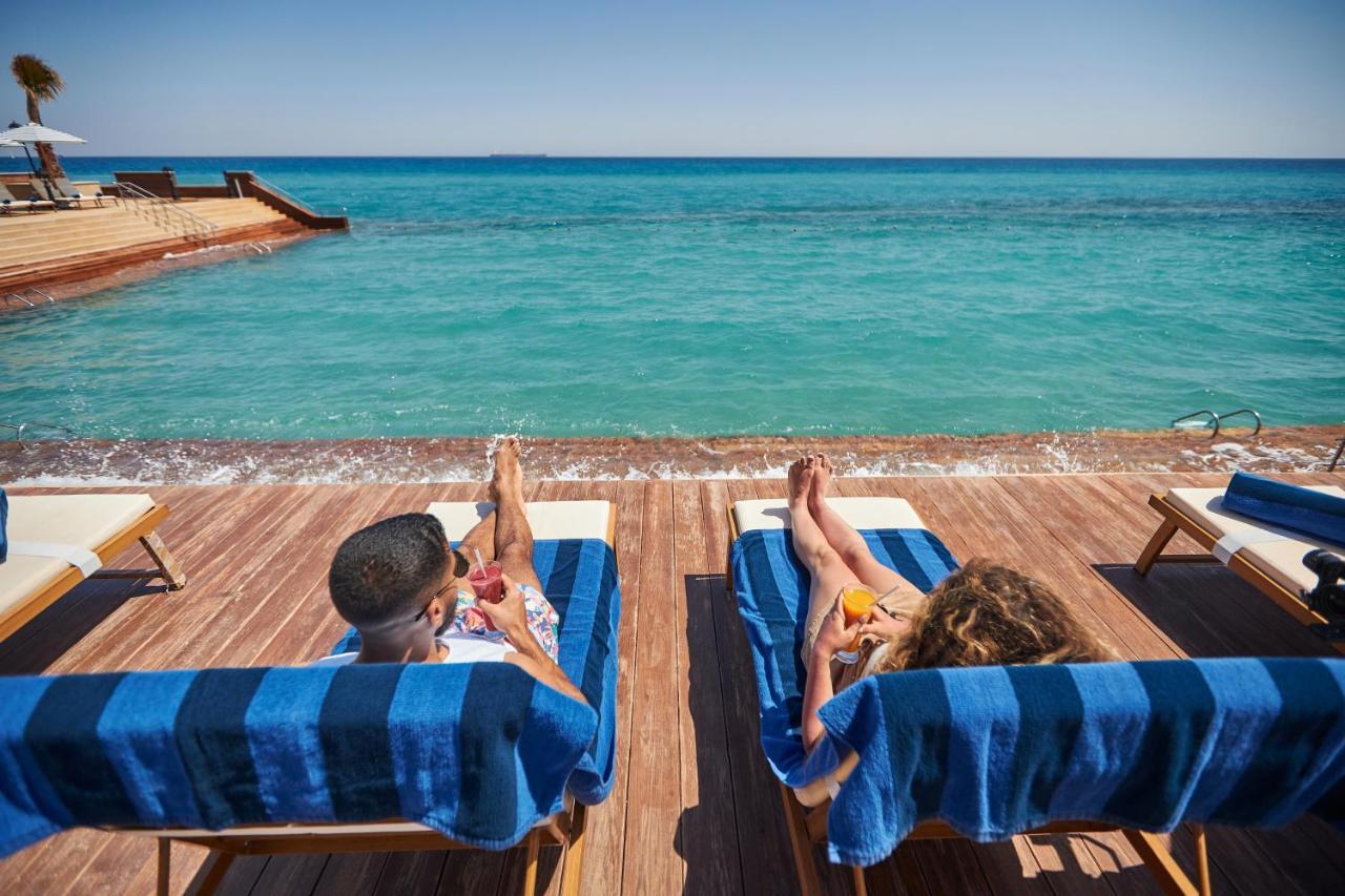 Tolip Resort El Galala Majestic Ain Sukhna Luaran gambar The photo depicts two people relaxing on lounge chairs by the waterfront. They are positioned on a wooden deck with a view of the clear, turquoise water. Both individuals are reclining, enjoying the sun, and each has a drink in hand. The scene convey