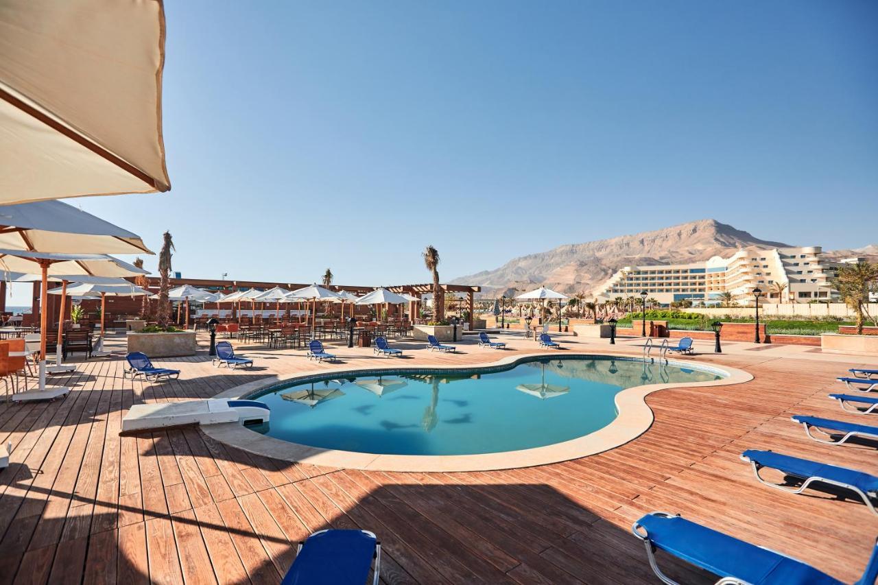 Tolip Resort El Galala Majestic Ain Sukhna Luaran gambar The photo shows a swimming pool area surrounded by sun loungers and umbrellas. The pool has a unique, curvy shape and is situated on a wooden deck. In the background, there are several buildings, likely part of a resort or hotel, and a mountainous la
