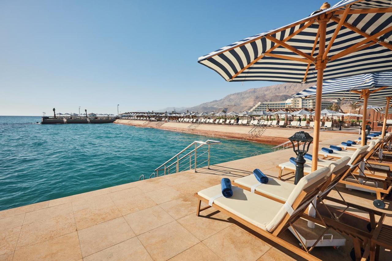 Tolip Resort El Galala Majestic Ain Sukhna Luaran gambar The photo shows a scenic beachside area with clear blue water. In the foreground, there are several sun loungers set up on a tiled surface, with blue towels neatly rolled on each one. The loungers are shaded by striped umbrellas. In the distance, a w