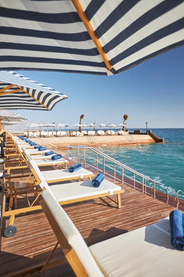 Tolip Resort El Galala Majestic Ain Sukhna Luaran gambar The photo depicts a serene outdoor area by the waterfront. There are several lounge chairs arranged in a row, each equipped with blue towels. Large, striped umbrellas provide shade over some of the chairs. In the background, there is a wooden deck th