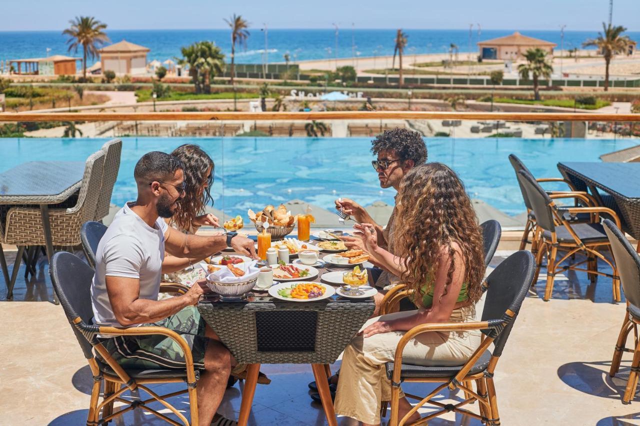 Tolip Resort El Galala Majestic Ain Sukhna Luaran gambar The photo shows a group of five people enjoying a meal together outdoors, likely at a restaurant or cafe. They are seated at a table with a beautiful view of a swimming pool and the sea in the background. The table is filled with various dishes, and 