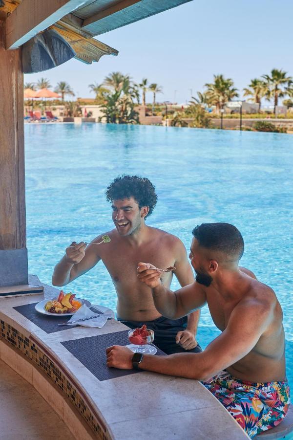Tolip Resort El Galala Majestic Ain Sukhna Luaran gambar The photo features two young men enjoying a meal together. They are sitting at a poolside table with a clear blue swimming pool in the background. The men are shirtless, suggesting a casual, relaxed atmosphere, likely in a warm environment. One man i