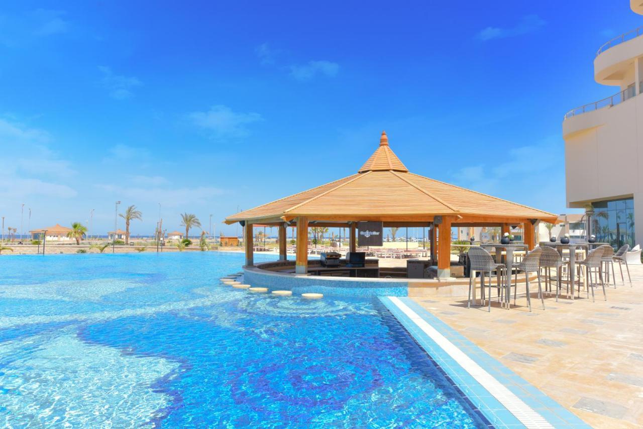 Tolip Resort El Galala Majestic Ain Sukhna Luaran gambar The photo shows a beautiful outdoor setting featuring a large swimming pool with clear blue water. In the center, there is a thatched-roof gazebo, which serves as a shaded seating area. Surrounding the gazebo are lounge chairs and a bar area with hig