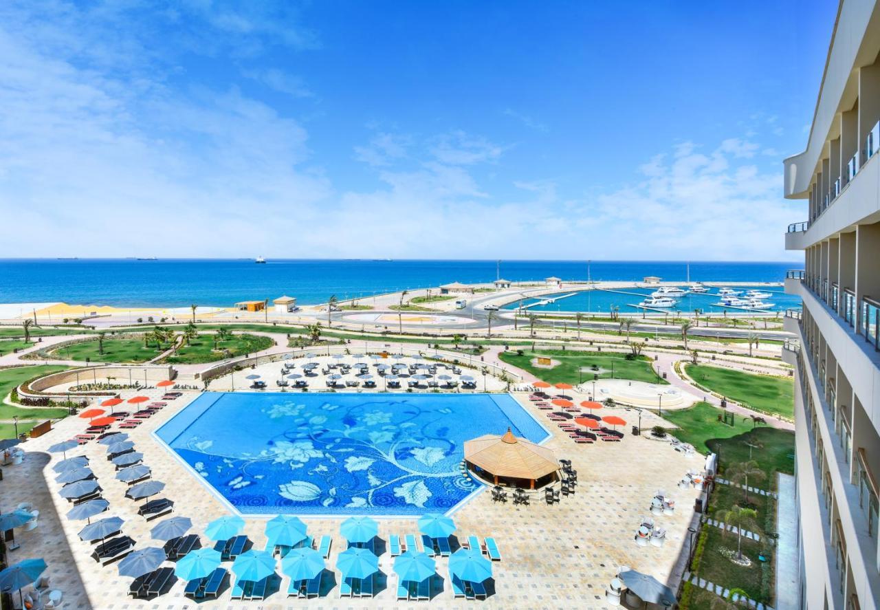 Tolip Resort El Galala Majestic Ain Sukhna Luaran gambar The photo shows a beautiful outdoor view of a resort area featuring a large swimming pool with a decorative underwater design. Surrounding the pool are numerous sun loungers and umbrellas in a variety of colors. In the background, there is a scenic c