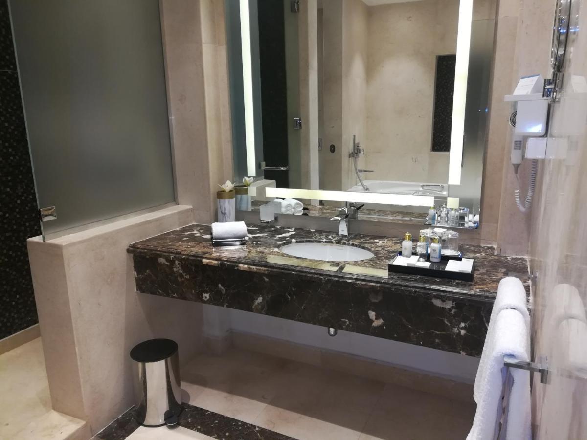 Tolip Resort El Galala Majestic Ain Sukhna Luaran gambar The photo shows a modern bathroom with elegant design features. There is a large vanity made of dark marble with a sink and a set of toiletries neatly arranged on top. A mirror with built-in lighting is mounted above the sink. To the side, there is a