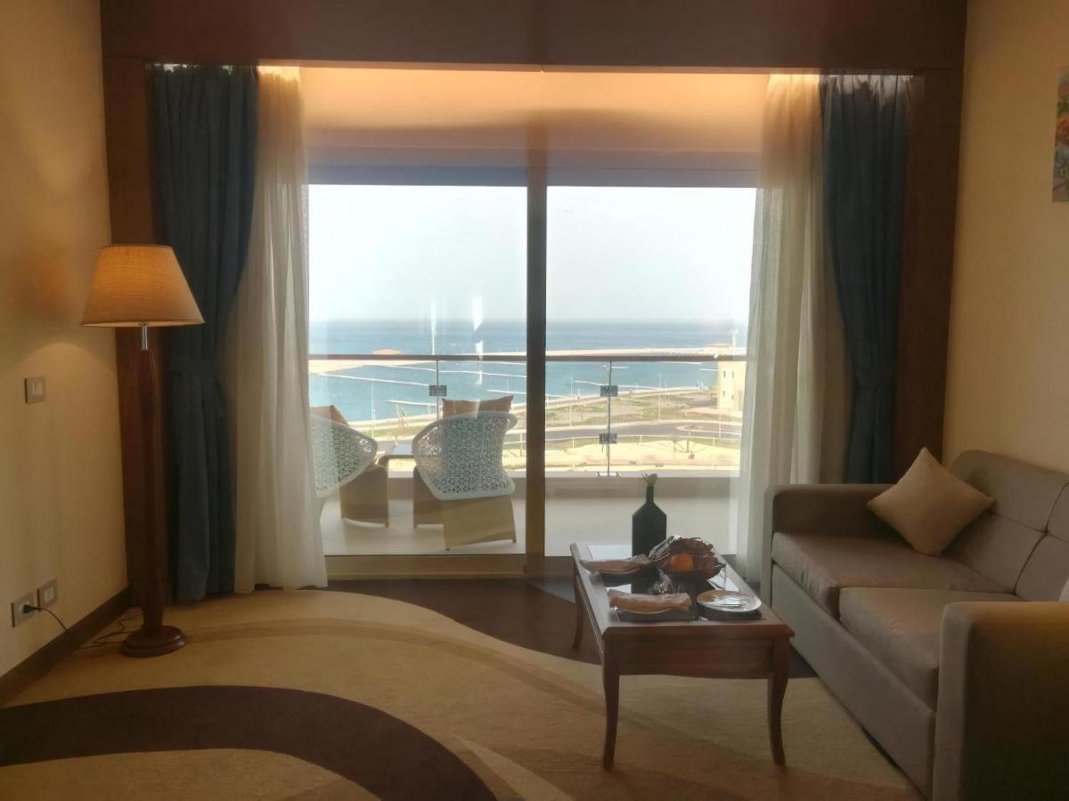 Tolip Resort El Galala Majestic Ain Sukhna Luaran gambar The photo shows a stylish interior of a room with a large window overlooking the sea. The window features sheer and thick curtains and is framed by a balcony with two chairs visible outside. Inside, there is a comfortable-looking sofa, a coffee table